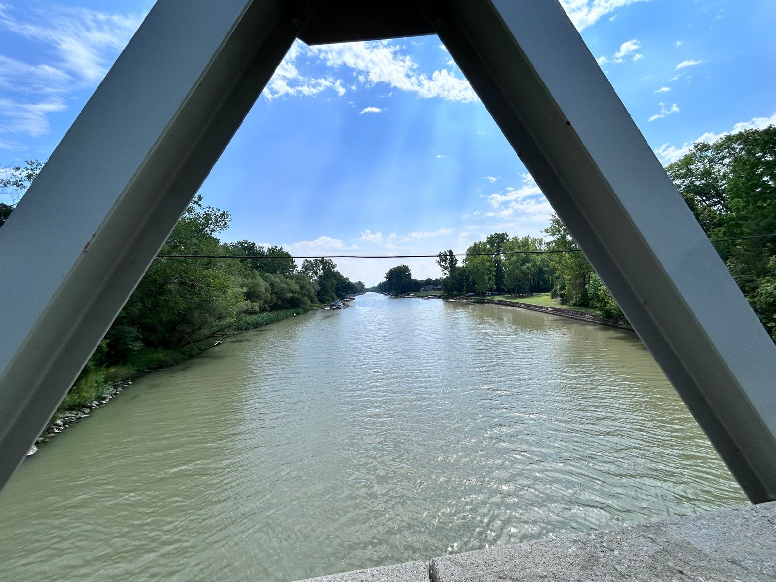 Looking out between the steel girders of a truss bridge along a wide river channel, moored boats and a stone seawall on the far bank