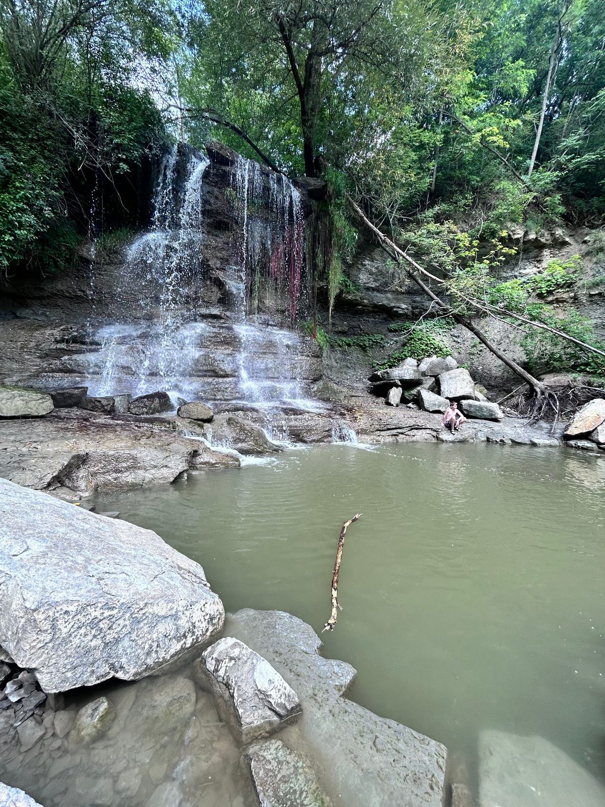 A waterfall spilling over a stepped face of grey shale into a shallow green plunge pool, a child sitting on the flat rock ledge at the right of the pool