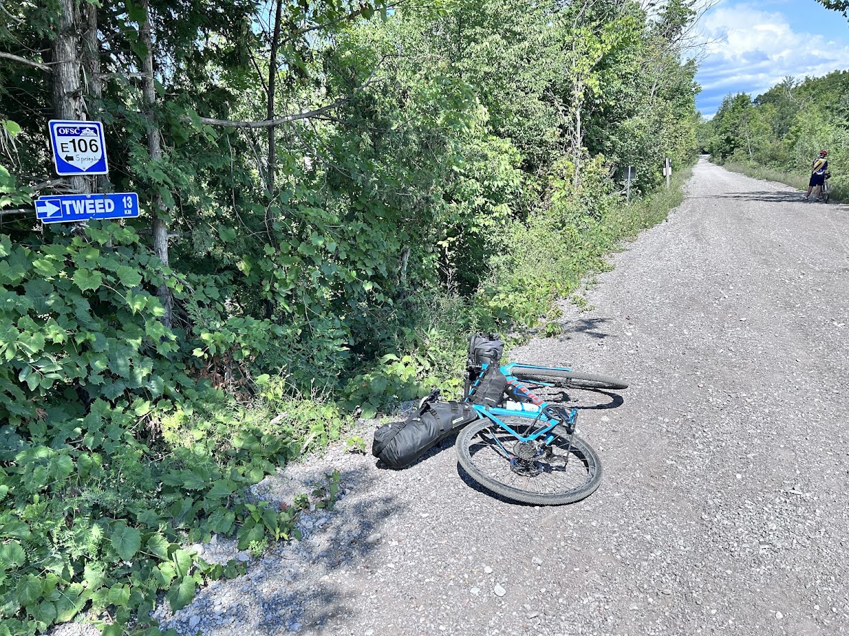 An OFSC trail marker reading Tweed 13 km, loaded gravel bike resting beside the trail